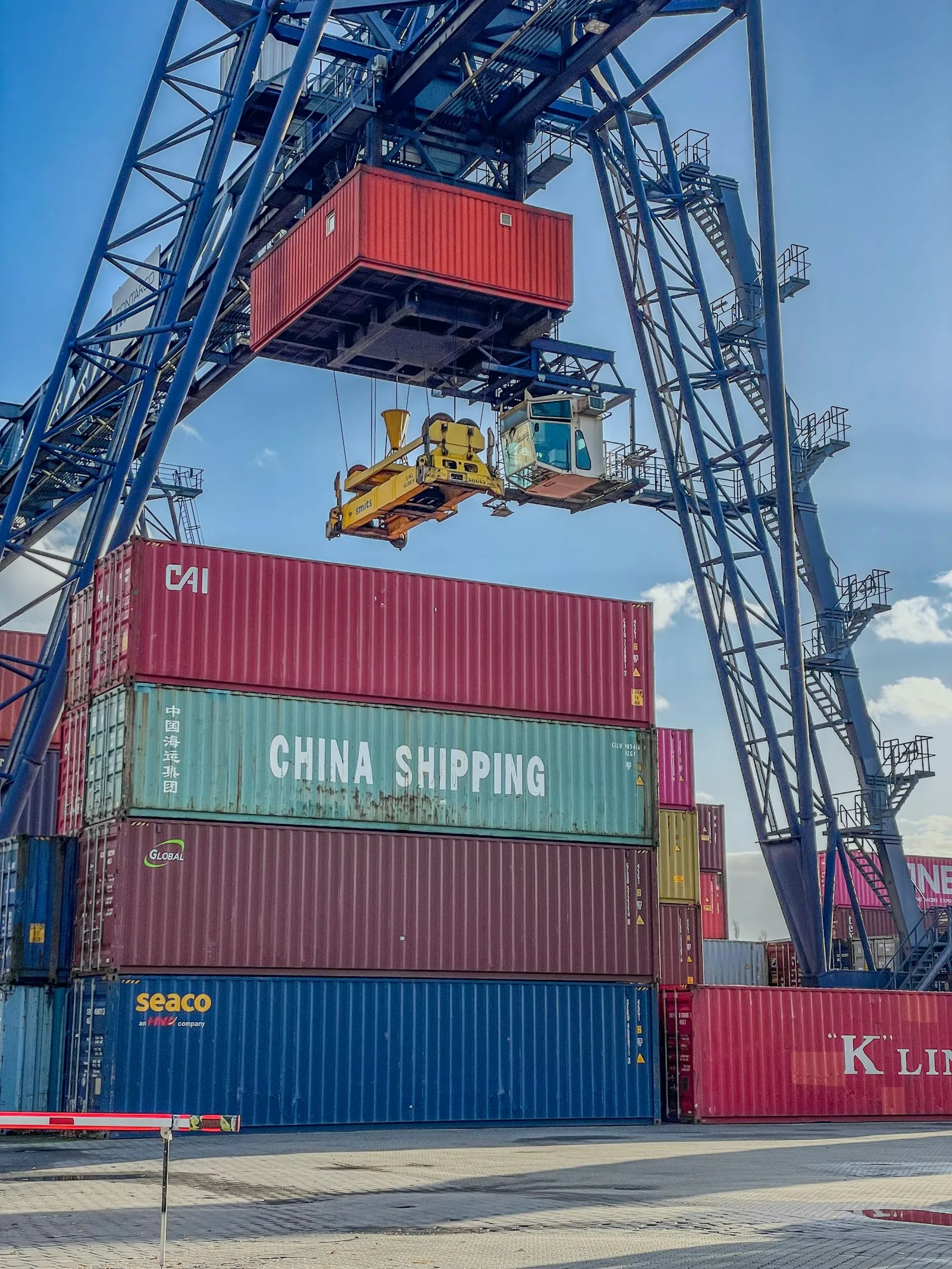 Containerterminal mit gestapelten Containern, darunter ein China-Shipping-Container, ein Kran hebt einen Container